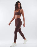 A woman modeling a matching brown activewear set consisting of a sports bra and high-waisted leggings, paired with white sneakers, against a light gray studio background.