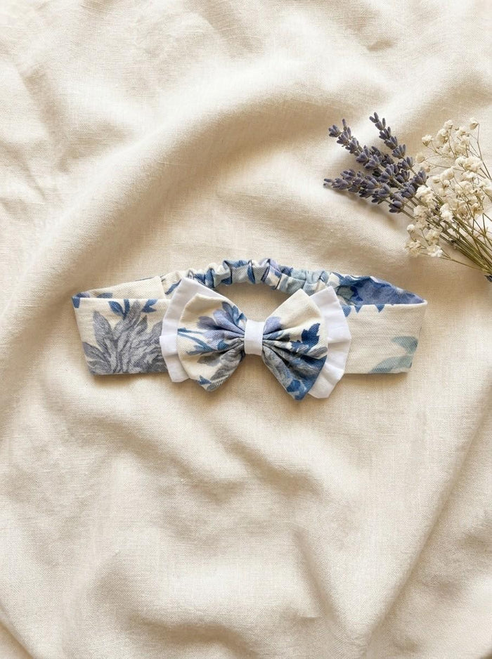 A flat lay photograph of the Alma baby headband, featuring a soft cream fabric with a blue and grey vintage-style floral pattern. The headband has an elasticated back for a comfortable fit and is topped with a matching floral bow that is layered over a white ruffled fabric. It is styled on a neutral, draped linen background next to a sprig of dried lavender and white baby's breath flowers.