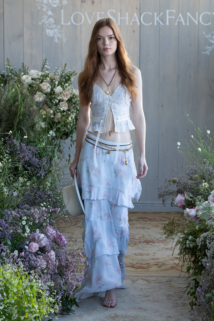 Woman in a floral tiered skirt and top standing in a garden with 'Love Shack Fancy' branding.