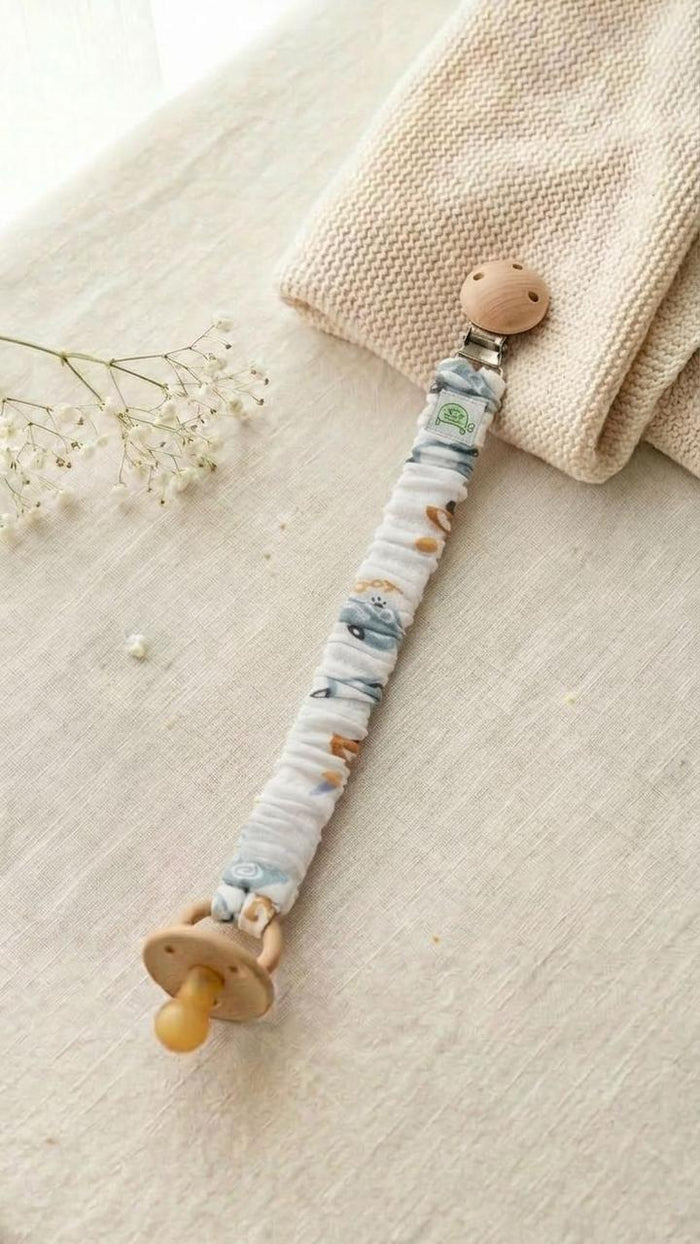 A flat lay photograph of a handmade fabric pacifier clip with a vintage car and vehicle print on a white background. The clip features a natural wooden fastener and holds a tan-colored pacifier. It is styled on a neutral linen surface next to a cream knitted blanket and a delicate sprig of white baby's breath flowers.
