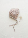 A flat lay photograph of the Carmen baby bonnet, featuring a neutral beige fabric with a dark brown vintage toile floral pattern. The bonnet has a ruffled brim, matching chin straps, and a small green brand label. It is styled on a crisp white linen background next to a delicate sprig of dried white baby's breath flowers.
