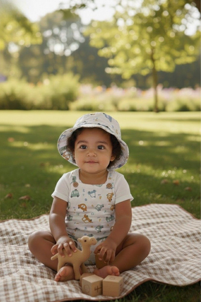 A lifestyle photograph of a young child sitting on a brown checkered picnic blanket in a sunny park. The child is wearing a white sleeveless romper and a matching bucket hat, both featuring a colorful print of cars and trucks. The child is playing with a wooden camel toy and wooden blocks, with a lush green lawn and trees in the soft-focus background.