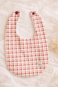 A flat lay photograph of the Cordoba baby bib, featuring a classic red and cream gingham checkered pattern. The bib is designed with a white textured, absorbent backing and two dark red snap fasteners at the neck. It is styled on a soft, wrinkled white linen background next to a small sprig of dried white flowers.