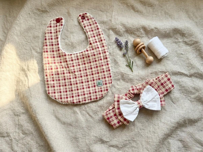 A flat lay photograph of the Cordoba set featuring a baby bib and a matching headband. Both items are made from a red and cream gingham checkered fabric. The bib has a white textured backing and red snap fasteners, while the headband features a layered double bow with a white eyelet fabric on top. They are styled on a neutral linen background alongside a wooden rattle, a sprig of dried lavender, and a small white cloth roll.