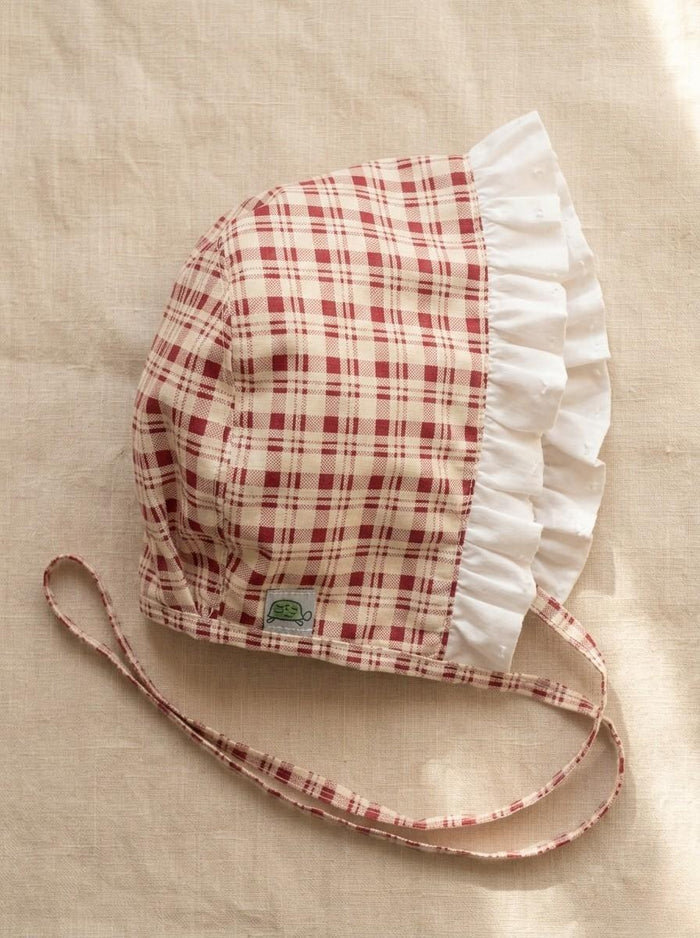 A flat lay photograph of the Cordoba baby bonnet, featuring a red-brown and cream checkered plaid fabric. The bonnet is designed with a wide, ruffled white fabric trim along the front brim and matching plaid chin straps. It is styled flat on a neutral, textured light beige linen background in soft, warm sunlight.