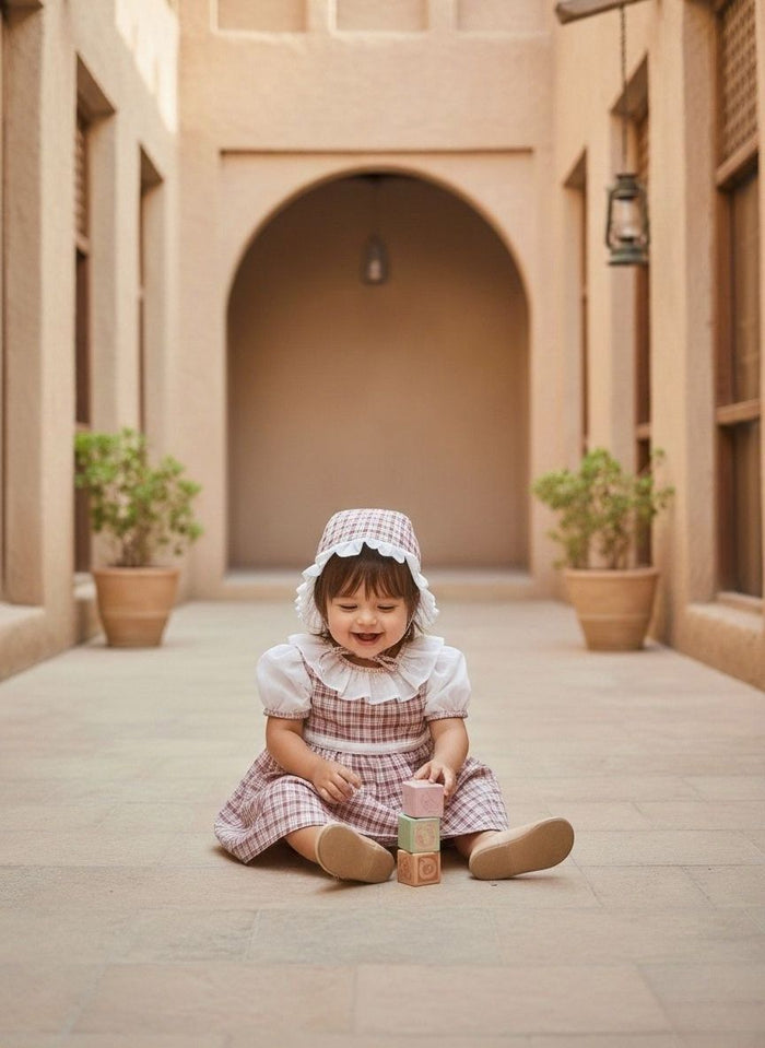 A lifestyle photograph of a smiling baby girl sitting on a stone-tiled floor in a warm, sandy-toned architectural corridor with a large archway in the background. She is wearing the Cordoba outfit, a short-sleeved dress and matching bonnet featuring a red-brown and cream plaid pattern. The dress is detailed with a white ruffled collar and white undersleeves, and the bonnet has a delicate white ruffled trim. She is playing with a small stack of pink and green toy blocks.