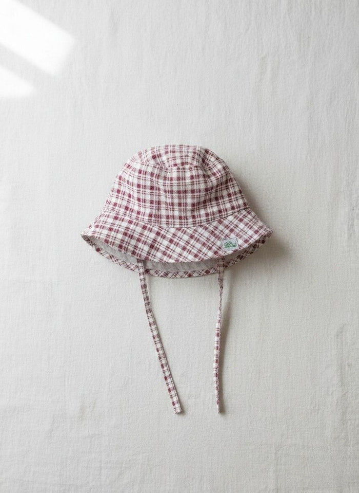 A top-down photograph of the Cordoba baby bucket hat, featuring a red-brown and cream checkered plaid fabric. The hat is designed with matching plaid chin tie straps and includes a small square green brand label stitched onto the side brim. It is styled flat and centered on a textured, light cream linen background in soft, natural light.