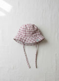 A top-down photograph of the Cordoba baby bucket hat, featuring a red-brown and cream checkered plaid fabric. The hat is designed with matching plaid chin tie straps and includes a small square green brand label stitched onto the side brim. It is styled flat and centered on a textured, light cream linen background in soft, natural light.