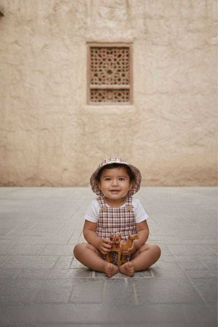 A portrait-style lifestyle photograph of a smiling baby sitting cross-legged and barefoot on a grey stone-tiled floor. The baby is wearing the Cordoba set, featuring short romper overalls and a matching bucket hat in a red-brown and cream checkered plaid fabric, styled over a plain white short-sleeved t-shirt. The baby is holding a small toy camel against a backdrop of a warm, sandy-coloured textured stone wall with an ornate lattice window cutout.