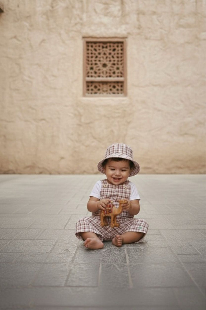 A lifestyle photograph of a smiling baby sitting barefoot on a grey stone-tiled floor. The baby is wearing the Cordoba set, which includes short romper overalls and a matching bucket hat in a red-brown and cream checkered plaid fabric, worn over a simple white short-sleeved t-shirt. The baby is holding a small toy camel against a backdrop of a warm, sandy-coloured textured stone wall with an ornate lattice window cutout.