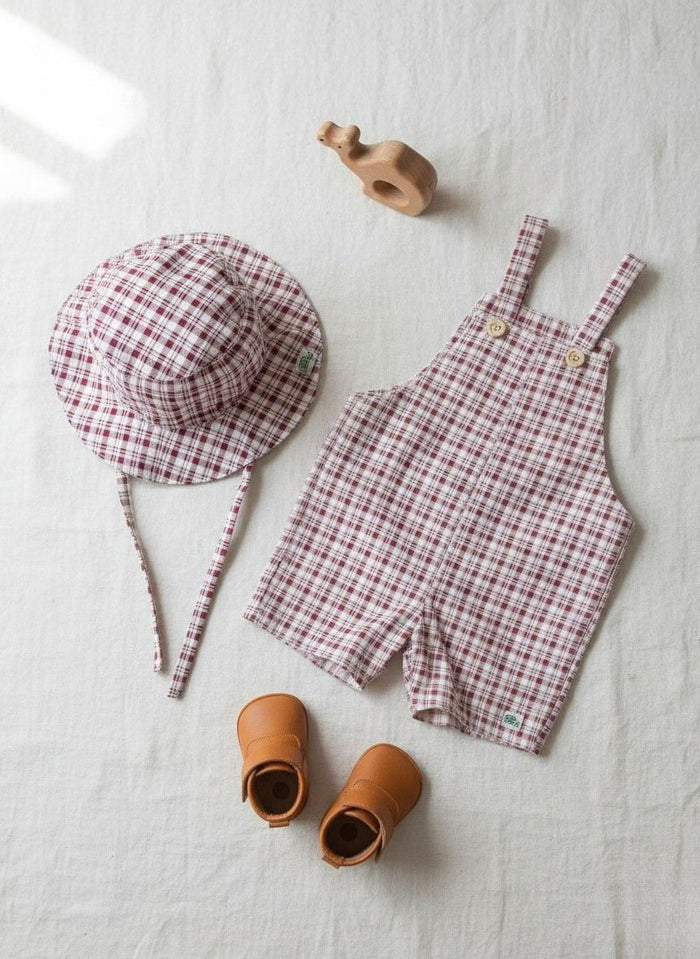A flat lay photograph of a baby outfit set featuring the Cordoba short romper overalls and a matching bucket hat. Both pieces are crafted from a red-brown and cream checkered plaid fabric. The overalls include small light wood buttons on the straps, and the bucket hat has matching fabric tie straps. The set is styled on a neutral cream linen background alongside a pair of tan leather baby booties and a small wooden animal toy.