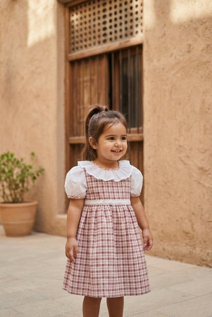 A portrait lifestyle photograph of a smiling young girl standing outside on a stone-tiled ground. She is wearing the Cordoba dress, which features a red-brown and cream checkered plaid print, white puffed short sleeves with plaid trim, a wide white ruffled collar, and a delicate white lace-style band across the waist. She has her hair styled in a high half-ponytail, against a background of a warm, sandy-toned stone wall with a rustic wooden window frame and a potted green plant.
