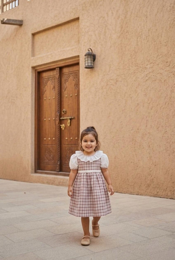 A lifestyle photograph of a smiling young girl walking forward on a stone-piled ground. She is wearing the Cordoba dress, featuring a red-brown and cream checkered plaid print, detailed with short puffed white sleeves, a wide ruffled white collar, and a delicate white waistband. She is wearing beige Mary Jane shoes against a background of a textured, sandy-brown stone building with a large, ornate vintage wooden double door and an old-fashioned wall lantern.