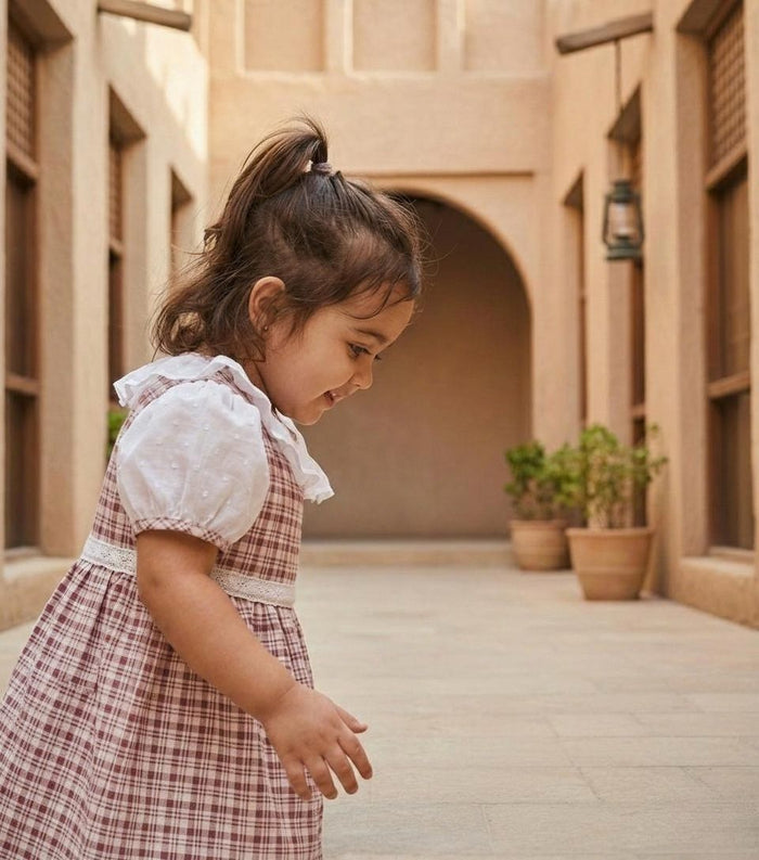 A side-angle lifestyle photograph of a young girl captured in motion while walking through a warm, sandy-toned stone corridor with high walls and potted plants in the background. She is wearing the Cordoba dress, featuring a classic red-brown and cream checkered plaid pattern. The design includes white puffed short sleeves with plaid trim, a white ruffled collar, and a delicate white lace-style band across the waistline. She has her hair pulled back into a half-ponytail and is looking downward w