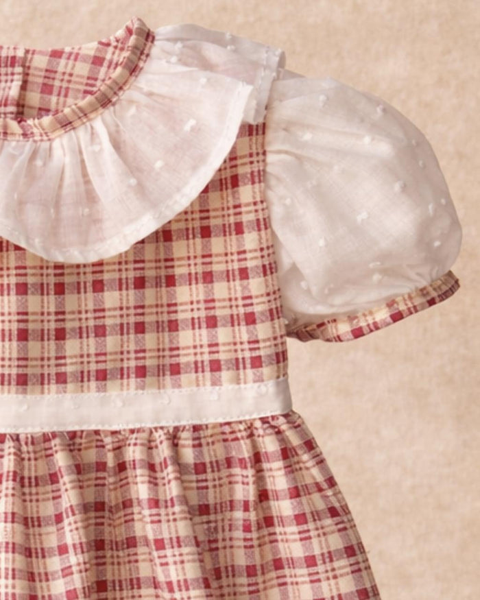 A close-up photograph focusing on the bodice and sleeve details of the Cordoba baby dress. The shot highlights the intricate red-brown and cream checkered plaid fabric paired with a wide, layered white ruffle collar made from a sheer swiss dot material. It shows a matching white puffed sleeve finished with a thin plaid cuff trim, and a delicate white swiss dot band detailing the waistline against a soft, blurred neutral background.