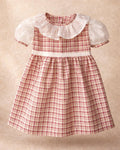 A flat lay photograph of the Cordoba baby dress, featuring a classic red-brown and cream checkered plaid pattern. The short-sleeved dress is designed with a wide, ruffled white collar made from sheer swiss dot fabric, matching puffed white undersleeves with plaid trim, and a thin white swiss dot waistband. It is displayed centered against a soft, neutral tan textured background.