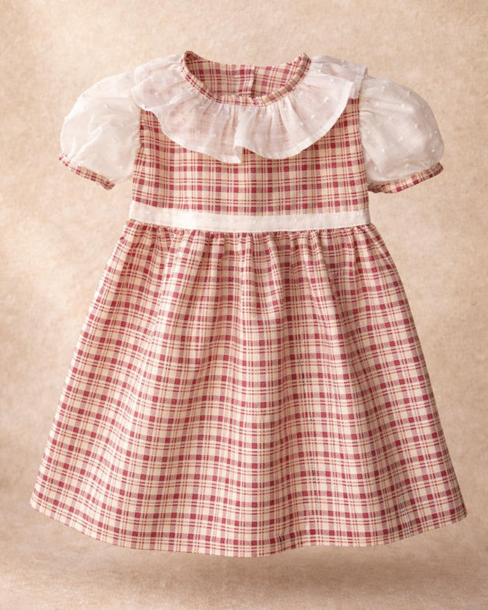 A flat lay photograph of the Cordoba baby dress, featuring a classic red-brown and cream checkered plaid pattern. The short-sleeved dress is designed with a wide, ruffled white collar made from sheer swiss dot fabric, matching puffed white undersleeves with plaid trim, and a thin white swiss dot waistband. It is displayed centered against a soft, neutral tan textured background.