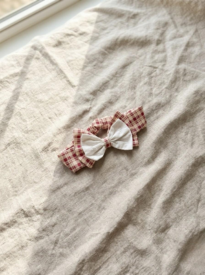 A flat lay photograph of the Cordoba baby headband featuring a red and cream gingham checkered pattern. The headband is topped with a unique double bow: a larger gingham bow sits beneath a smaller, delicate white eyelet fabric bow. It is styled diagonally on a textured, neutral-toned linen fabric under soft, natural sunlight.