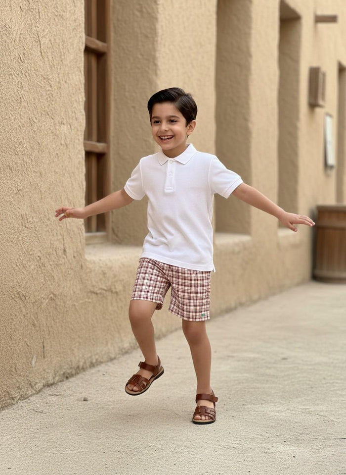 An action lifestyle photograph of a laughing young boy skipping playfully outdoors in an alley with rustic, sandy-toned stone walls. He is wearing the Cordoba outfit, which features a clean white short-sleeved polo shirt paired with red-brown and cream checkered plaid shorts. He has his arms outstretched for balance and is wearing brown leather strappy sandals.