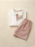 A flat lay photograph of a little boy's clothing set featuring a folded white short-sleeved polo shirt and matching plaid shorts. The polo shirt includes a collar and a button placket lined with a red-brown and cream checkered fabric. The coordinating shorts feature an elastic waistband in the same red-brown plaid print and a small green turtle brand label at the hem, all styled on a neutral, cream-colored fabric background.