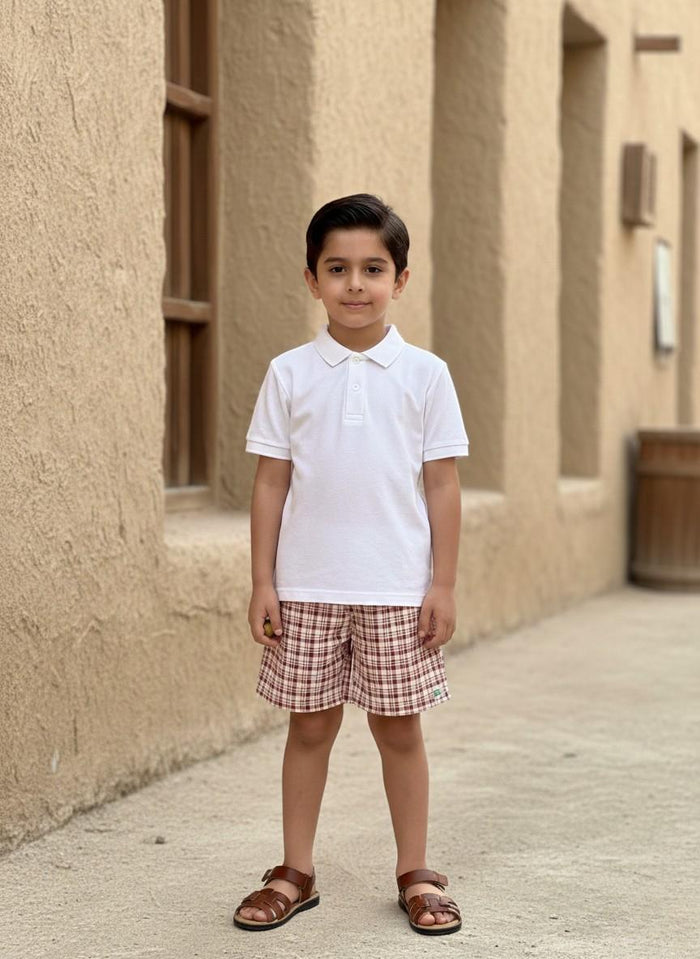 A lifestyle photograph of a young boy standing outside in a historic-looking alley with textured, sandy-brown stone walls. He is wearing the Cordoba set, which consists of a clean white short-sleeved polo shirt and matching red-brown and cream plaid shorts. The outfit is paired with brown leather strappy sandals.
