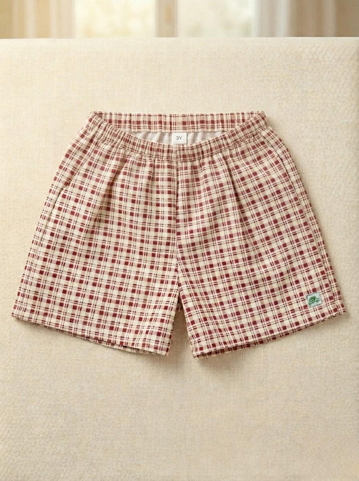 A flat lay photograph of the Cordoba boys' shorts, featuring a red-brown and cream checkered plaid pattern. The shorts are designed with a comfortable elastic waistband and include a small green square brand label near the bottom left hem. They are displayed flat on a neutral, textured light cream fabric background.