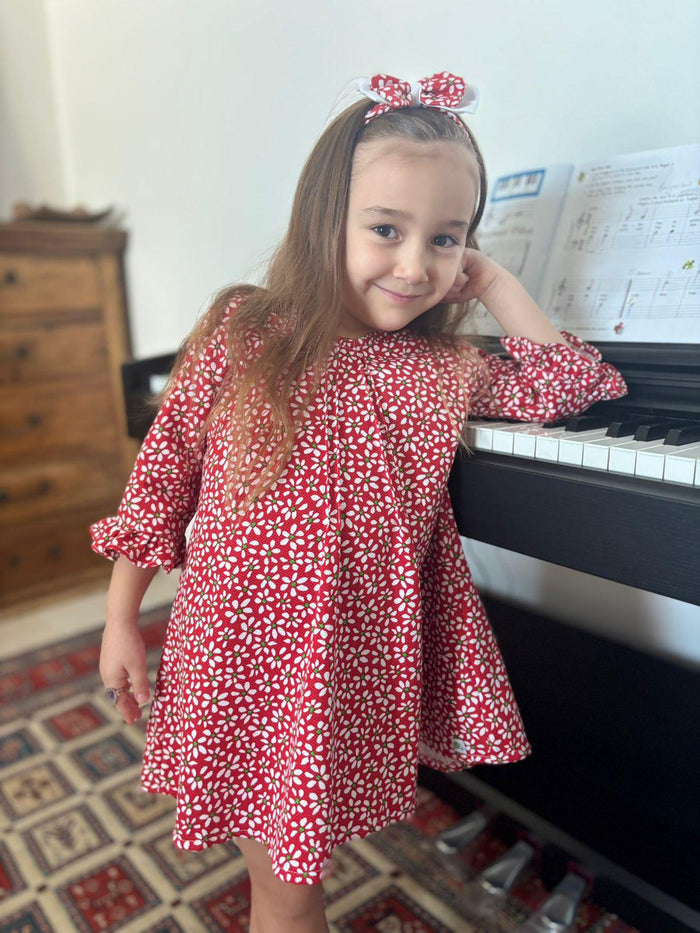 A lifestyle photograph of a young girl with long brown hair smiling while leaning against a black piano. She is wearing the Elena dress, a vibrant red A-line dress with a white daisy floral pattern and ruffled long sleeves. She is also wearing the matching Elena floral headband.