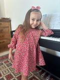 A lifestyle photograph of a young girl with long brown hair smiling while leaning against a black piano. She is wearing the Elena dress, a vibrant red A-line dress with a white daisy floral pattern and ruffled long sleeves. She is also wearing the matching Elena floral headband.