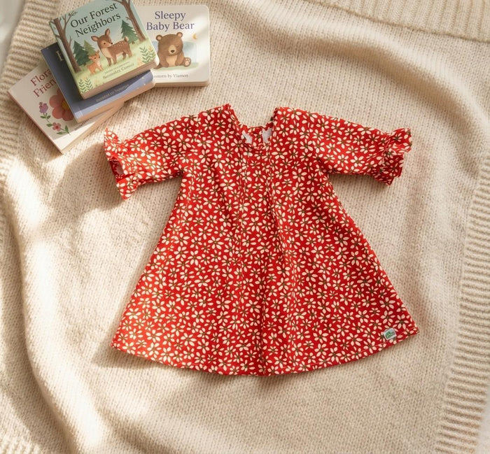 A flat lay photograph of the Elena baby dress in a vibrant red with a white daisy floral print. The dress features a classic A-line silhouette, ruffled sleeve cuffs, and a small green turtle brand label - la Tortuga Marisa label - at the hem. It is styled on a cream-colored knitted blanket alongside a small stack of children's board books.
