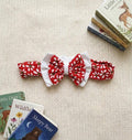 A flat lay photograph of the Elena baby headband, featuring a wide red band with a white floral daisy pattern. The headband is adorned with a large double-layered bow—a white fabric base topped with a matching red floral bow. It is styled on a cream knitted background, surrounded by several children's board books.