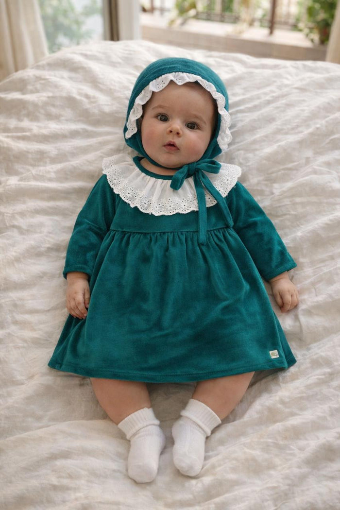 A lifestyle photograph of a baby girl lying on a soft white bed, wearing an emerald green velvet dress with a white eyelet lace collar and a matching velvet bonnet with lace trim. The baby is also wearing white socks, and the setting is bright and airy.