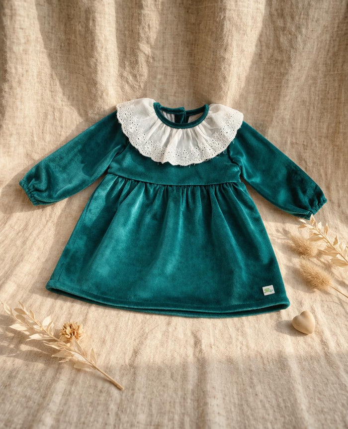 A long-sleeved emerald green velvet baby dress featuring a wide white eyelet lace Peter Pan collar. The dress has a gathered waist and elasticated cuffs, displayed on a neutral linen backdrop with dried botanical accents and a small wooden heart.
