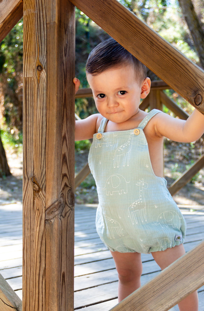 A portrait lifestyle photograph of a young baby boy standing on an outdoor wooden deck. He is wearing a mint green or light sage bubble romper featuring a white line art pattern of giraffes and elephants, detailed with small wooden buttons on the shoulder straps. The baby is looking directly at the camera with a gentle expression while peeking through a rustic wooden railing structure.
