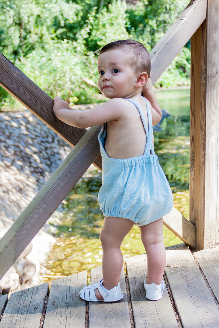 A lifestyle photograph of a baby boy standing on a rustic wooden bridge or dock. He is wearing a light blue bubble-style romper featuring a subtle giraffe print, designed with crossed shoulder straps and an elastic back waistband. The baby is wearing white leather closed-toe sandals and looks back over his shoulder while holding onto a wooden railing. The background features a sunny outdoor setting with a riverbank and lush green trees.