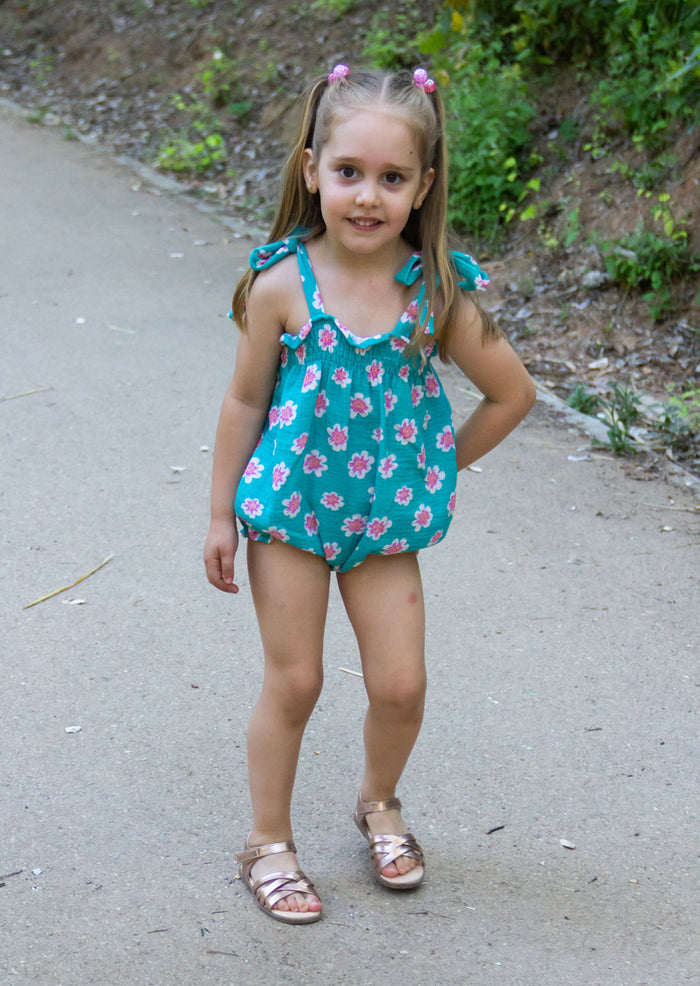 A full-length portrait lifestyle photograph of a smiling young blonde girl standing on a concrete path. She is wearing the Joy romper, a bright teal bubble-style romper featuring a print of pink and white flowers, tie-shoulder straps, and a smocked bodice. Her hair is styled in two pigtails on top secured with pink hair ties, and she is wearing rose gold metallic strappy sandals while posing with one hand toward her hip against a background with a sloped grassy edge.