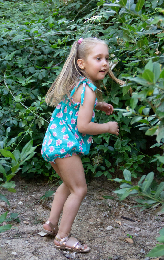 A full-length lifestyle photograph of a young blonde girl captured in a playful, dynamic stance outdoors. She is wearing the Joy romper, a teal bubble-style romper featuring a pink and white floral pattern, shoulder straps tied into bows, and a smocked bodice. She is styled with her hair in a half-up ponytail secured by a pink hair tie and is wearing metallic rose gold strappy sandals. She stands on a dirt path, leaning forward as if exploring, with a dense wall of lush green leafy bushes behind