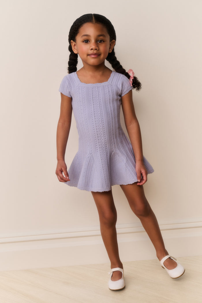 Young girl wearing a light blue dress with square neckline standing against a plain background