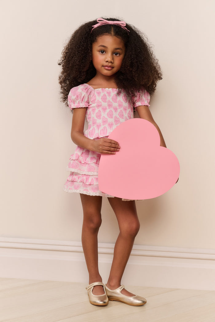 Young girl in a pink dress holding a heart-shaped object against a plain background