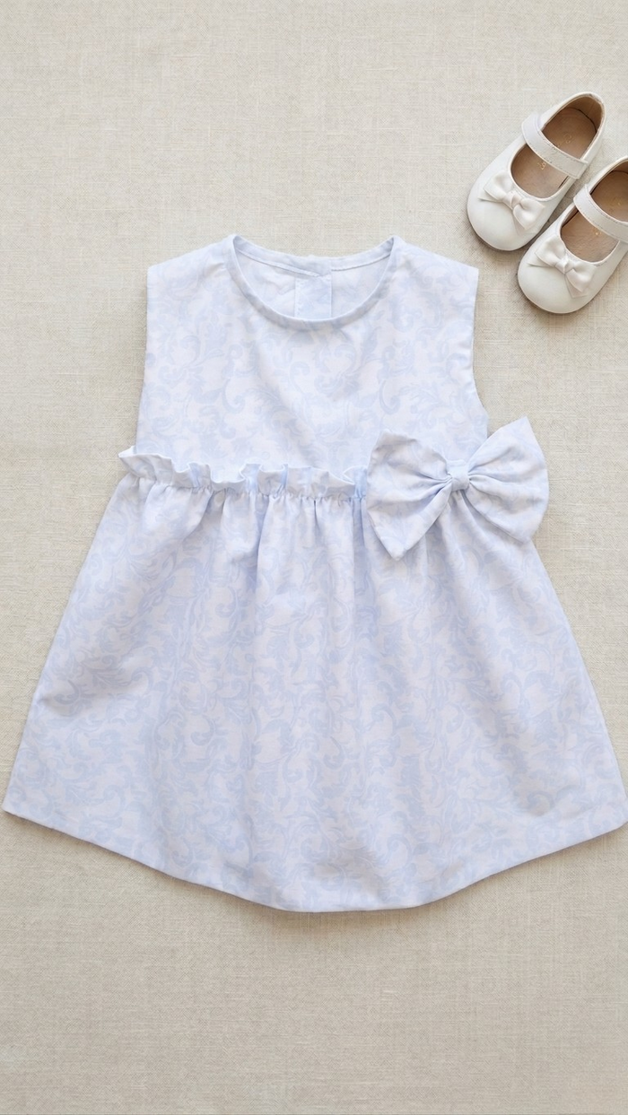 A flat lay photograph of the Malaga sleeveless baby dress in a light blue and white damask-style swirl pattern. The dress features a ruffled empire waistline with a large matching fabric bow on the side. It is styled on a neutral linen background next to a pair of small white Mary Jane shoes with matching bows.