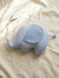 A handmade elephant-shaped plush cushion featuring a blue and white vertical striped body and a solid light blue muslin fabric ear. The cushion is displayed on a neutral linen backdrop with a sprig of dried lavender in the upper left corner, creating a soft and calm nursery aesthetic.