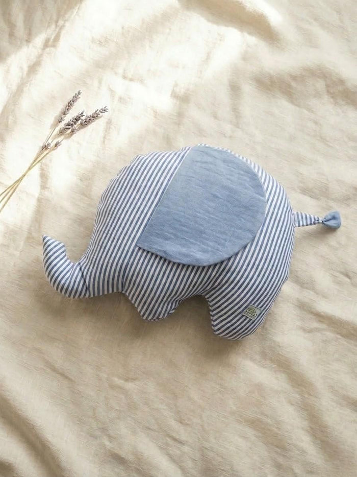 A handmade elephant-shaped plush cushion featuring a blue and white vertical striped body and a solid light blue muslin fabric ear. The cushion is displayed on a neutral linen backdrop with a sprig of dried lavender in the upper left corner, creating a soft and calm nursery aesthetic.