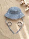 A top-down photograph of the Menorca baby bucket hat, featuring a light blue and white fine pinstriped fabric adorned with small embroidered white daisies. The hat includes matching pinstriped chin tie straps and a small square turtle brand label on the side. It is styled flat on a textured, neutral beige linen background alongside a natural cotton boll branch under soft sunlight.