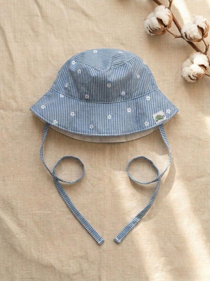 A top-down photograph of the Menorca baby bucket hat, featuring a light blue and white fine pinstriped fabric adorned with small embroidered white daisies. The hat includes matching pinstriped chin tie straps and a small square turtle brand label on the side. It is styled flat on a textured, neutral beige linen background alongside a natural cotton boll branch under soft sunlight.