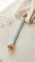 A flat lay photograph of a handmade baby pacifier clip in a soft mint green waffle-weave fabric. The clip features a natural round wooden fastener with a small green turtle brand label and holds a tan-colored pacifier. It is styled on a neutral linen surface next to a folded cream knitted blanket and a delicate sprig of white baby's breath flowers.