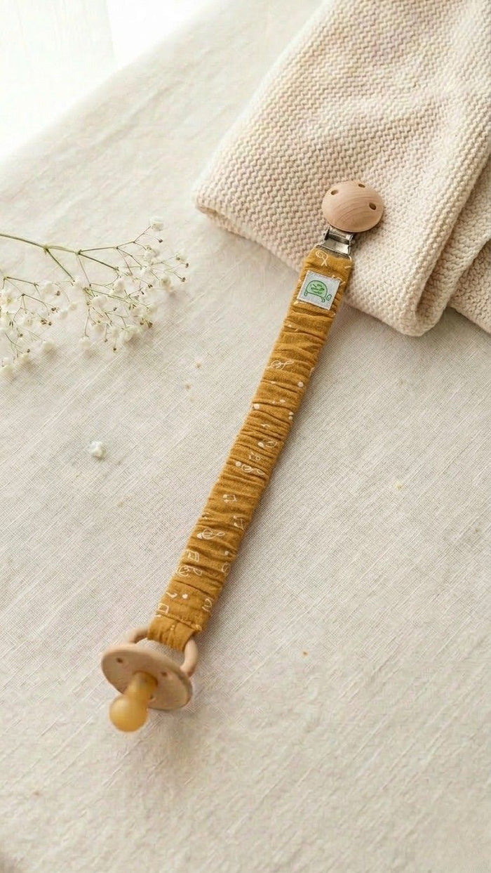 A flat lay photograph of a handmade fabric pacifier clip in mustard yellow, featuring a subtle white music note and staff print. The clip has a natural round wooden fastener at one end and holds a tan-colored pacifier at the other. It is styled on a neutral linen surface next to a folded cream knitted blanket and a sprig of white flowers.