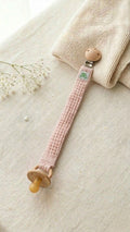 A flat lay photograph of a handmade baby pacifier clip in a soft dusty pink waffle-weave fabric. The clip features a round natural wooden fastener with a small green turtle brand label and holds a tan-colored pacifier. It is styled on a neutral linen surface next to a folded cream knitted blanket and a delicate sprig of white baby's breath flowers.