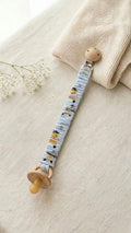A flat lay photograph of a handmade fabric pacifier clip featuring a pirate-themed print with anchors and treasure map elements on a white background. The clip includes a light brown wooden fastener and a matching blue loop for the pacifier. It is styled on a neutral linen fabric alongside a sprig of dried lavender and a small smooth stone.