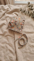 A side-view photograph of the Primavera baby bonnet, featuring a sage green fabric with a delicate pink and orange floral pattern. The bonnet includes a matching ruffled trim around the face and long fabric ties. It is styled on a neutral linen backdrop with a sprig of eucalyptus in the upper corner.