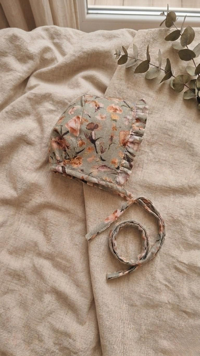 A side-view photograph of the Primavera baby bonnet, featuring a sage green fabric with a delicate pink and orange floral pattern. The bonnet includes a matching ruffled trim around the face and long fabric ties. It is styled on a neutral linen backdrop with a sprig of eucalyptus in the upper corner.