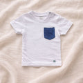 A flat lay photograph of a white, short-sleeved baby t-shirt featuring a single chest pocket made from dark blue velvet fabric. The shirt includes two silver snap fasteners on the left shoulder for easy dressing and a small green turtle brand label at the bottom right hem. It is styled against a soft, draped cream-colored linen background.